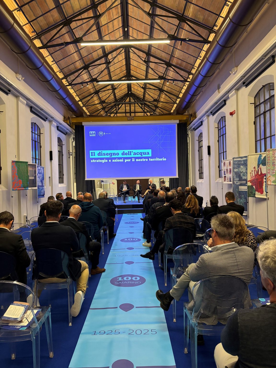 Sala conferenze con pubblico, proiezione centrale del logo “100 Saiarino” e grafica dell’acqua; manifesti artistici del territorio alle pareti