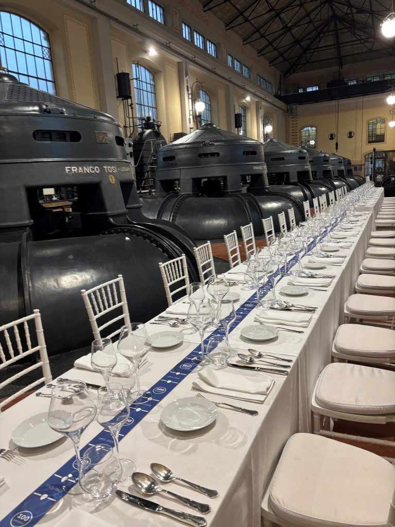 Tavola allestita con piatti e posate, fascia blu con logo “100 Saiarino” e strutture metalliche dell’impianto idrovoro sullo sfondo